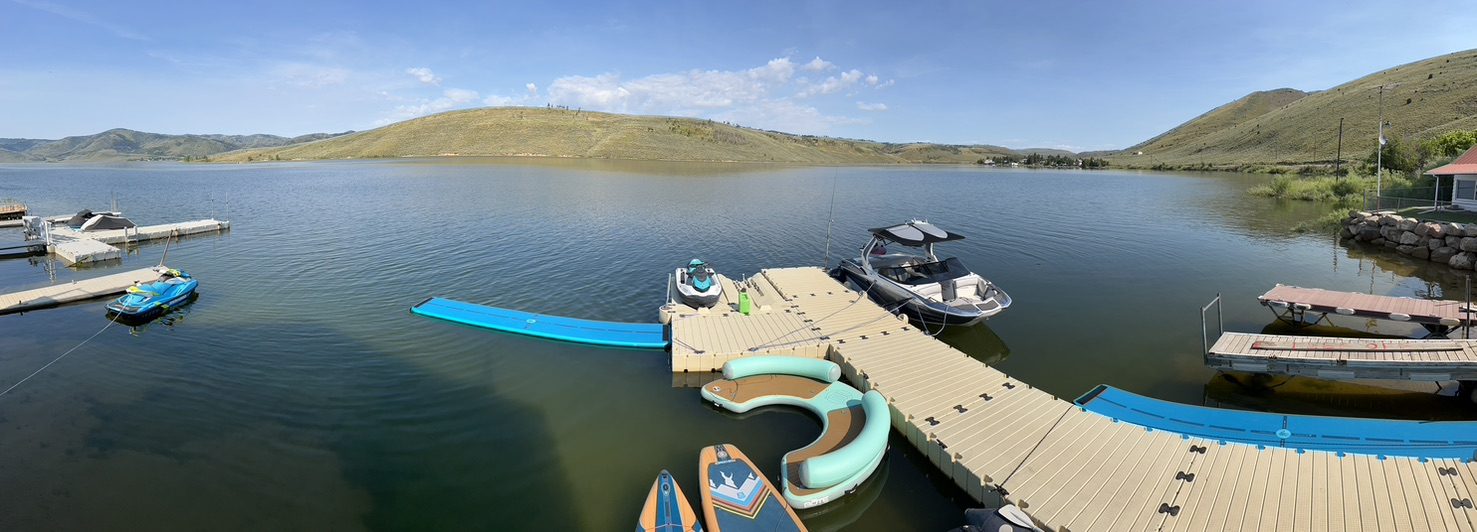 Panoramic view of the private dock on Scofield Reservoir with a wake boat, jet skis, paddleboards and a floating lounge, hills rising across the water